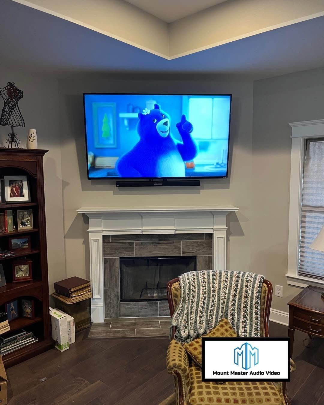 Living room with TV above fireplace; blue cartoon on screen, armchair, bookshelf, and window.