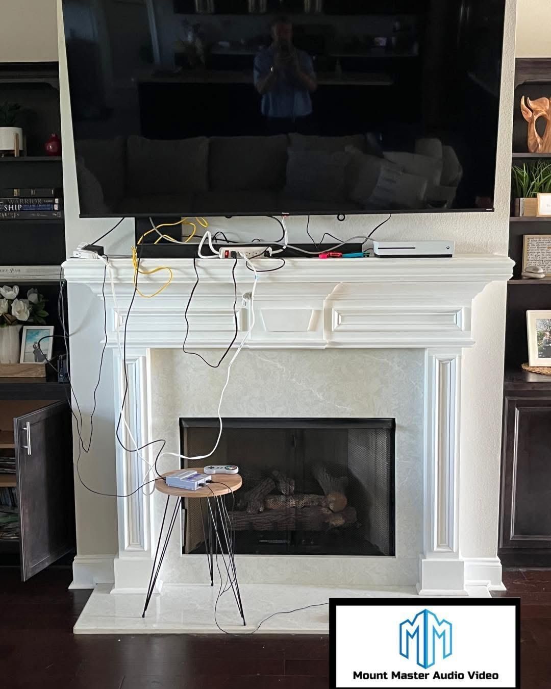 A fireplace with a TV mounted above it. Many wires are visible. A person is reflected in the TV.