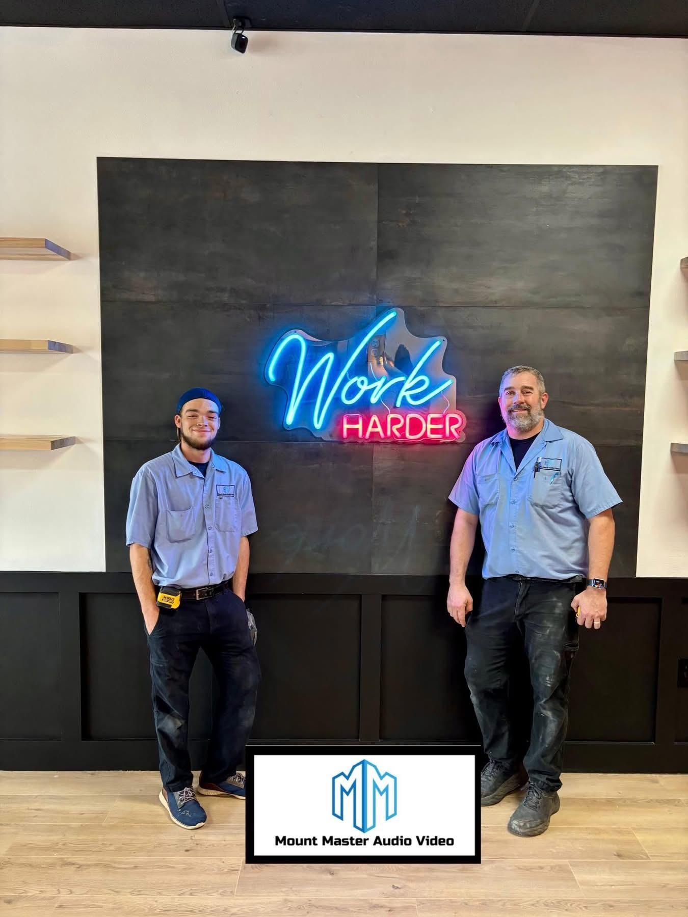 Two men in work uniforms stand in front of a wall with a neon