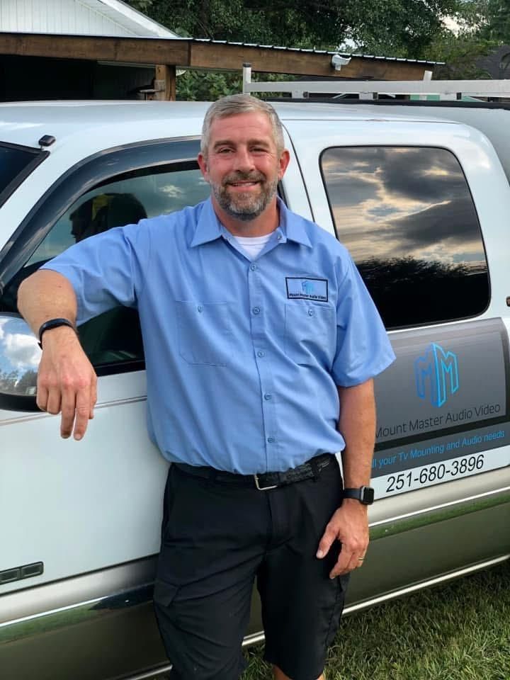 Man in blue work shirt and black shorts leaning against a service truck; smiles at the camera.