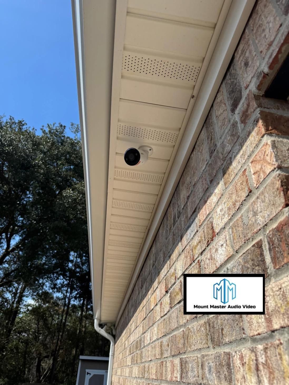 Security camera mounted on beige soffit next to a brick wall. Clear blue sky in the background.