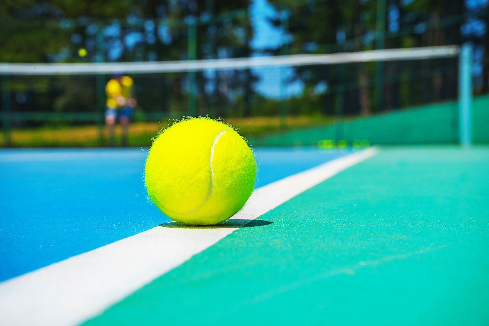 A bright yellow tennis ball resting on the white boundary line of a blue and green tennis court.