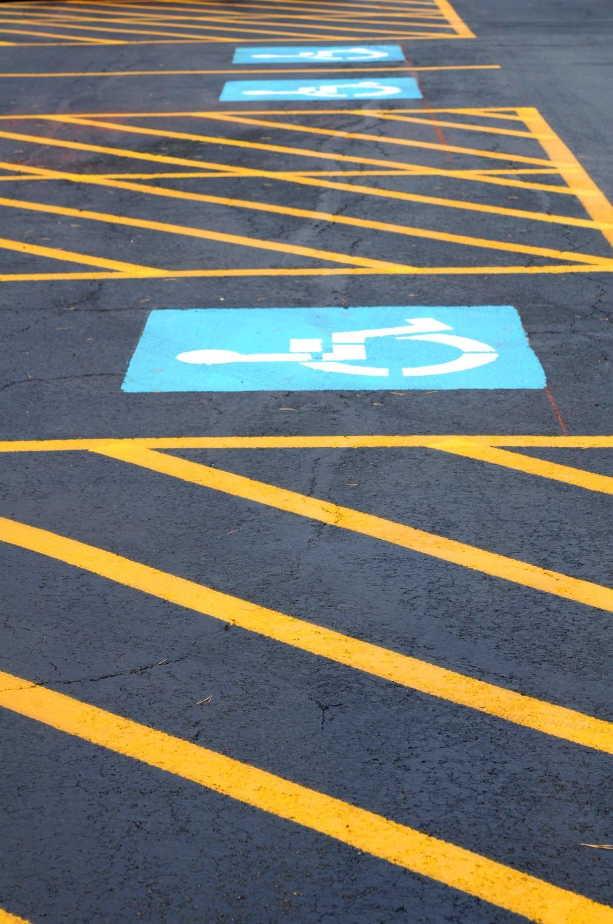 A parking lot asphalt surface with painted blue handicap symbols and yellow diagonal safety stripes.