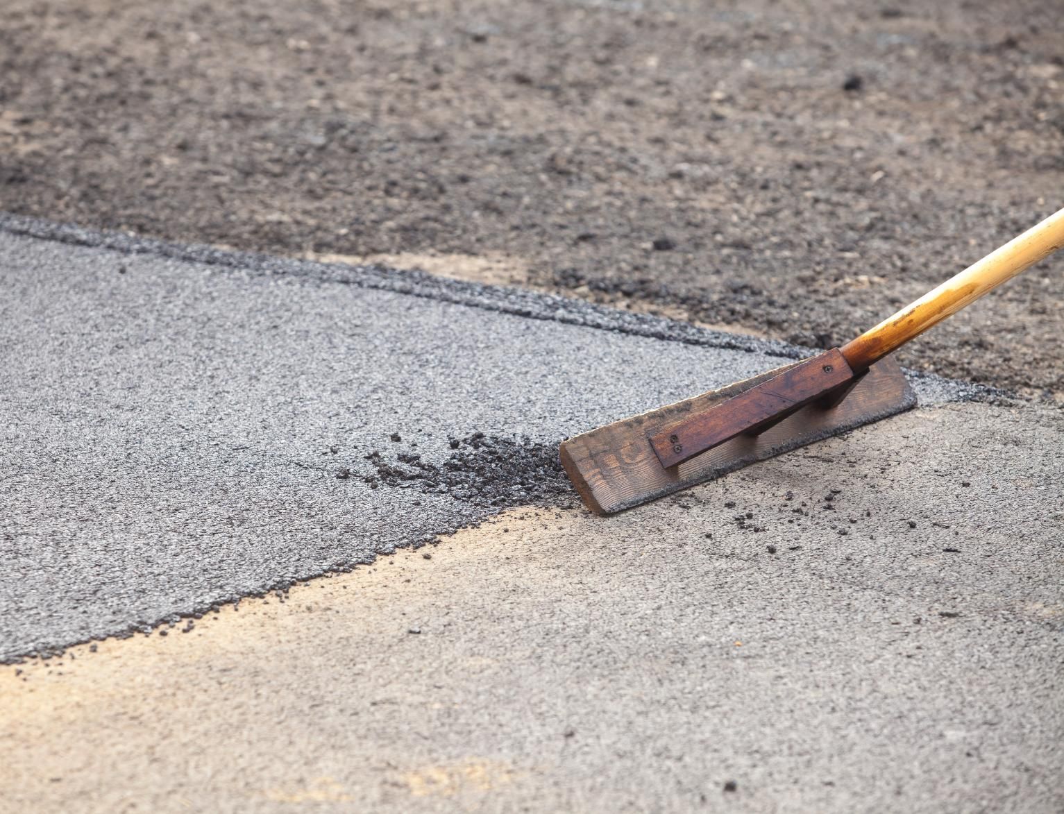 A wooden lute tool levels fresh, dark asphalt on a road surface being repaired.