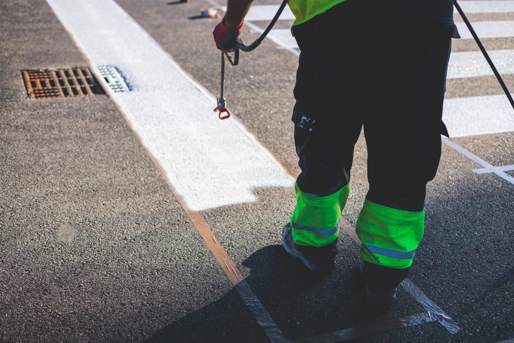 A worker in high-visibility neon leg gear paints white traffic stripes on an asphalt road near a drainage grate.