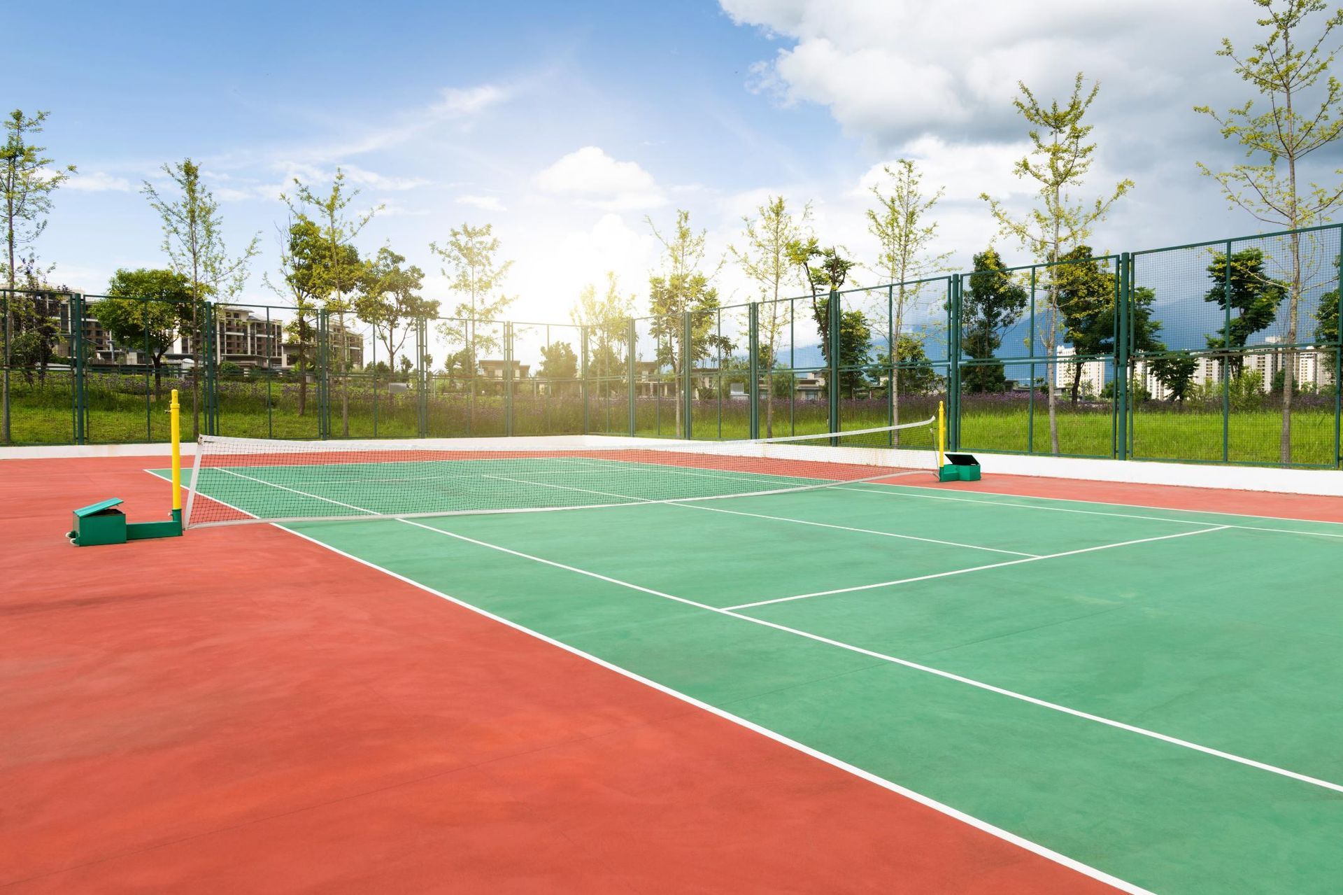 A tennis court with green and red flooring, a net, and a backdrop of green trees under a bright sky.