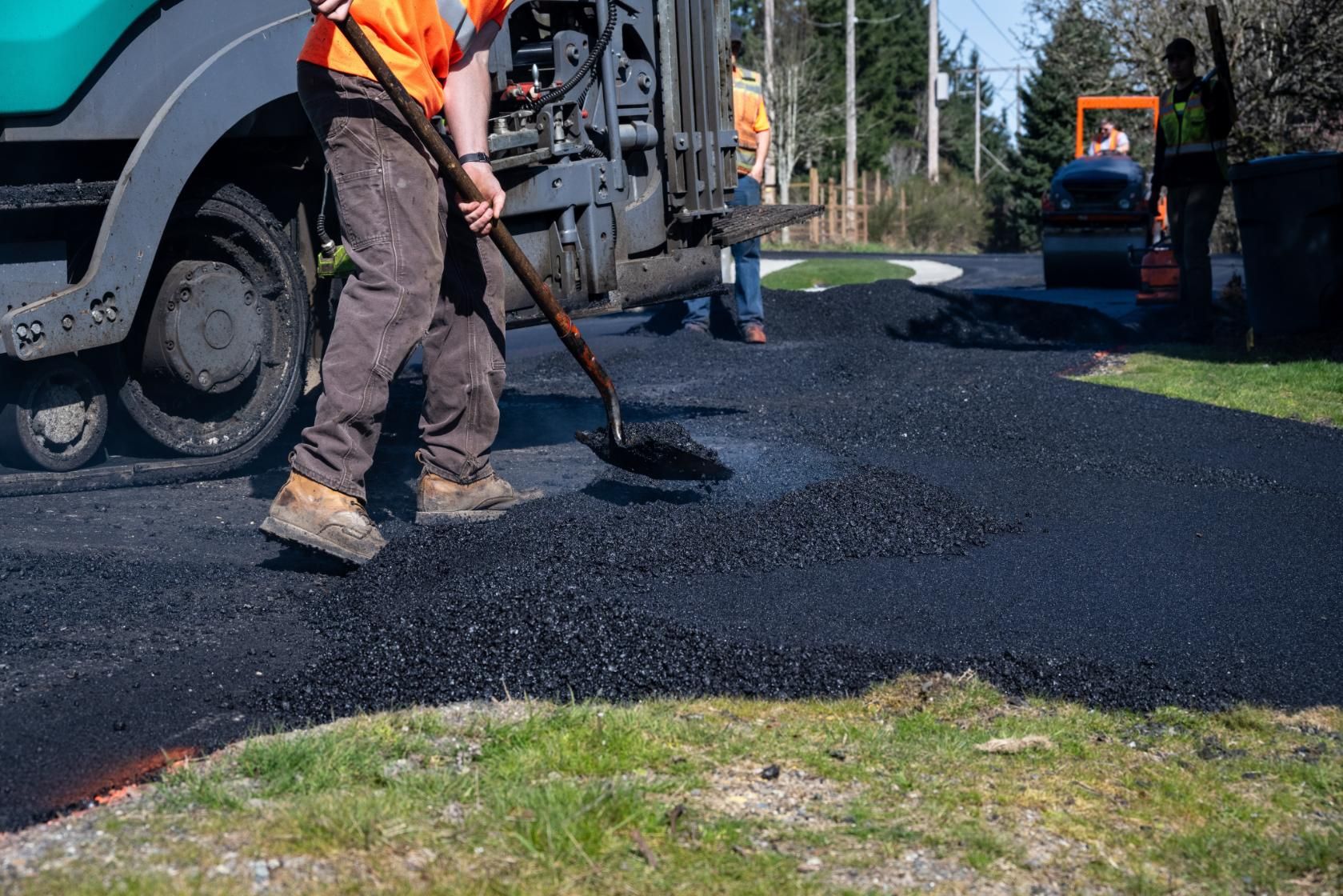 Workers use a shovel and heavy machinery to spread fresh asphalt on a residential driveway.