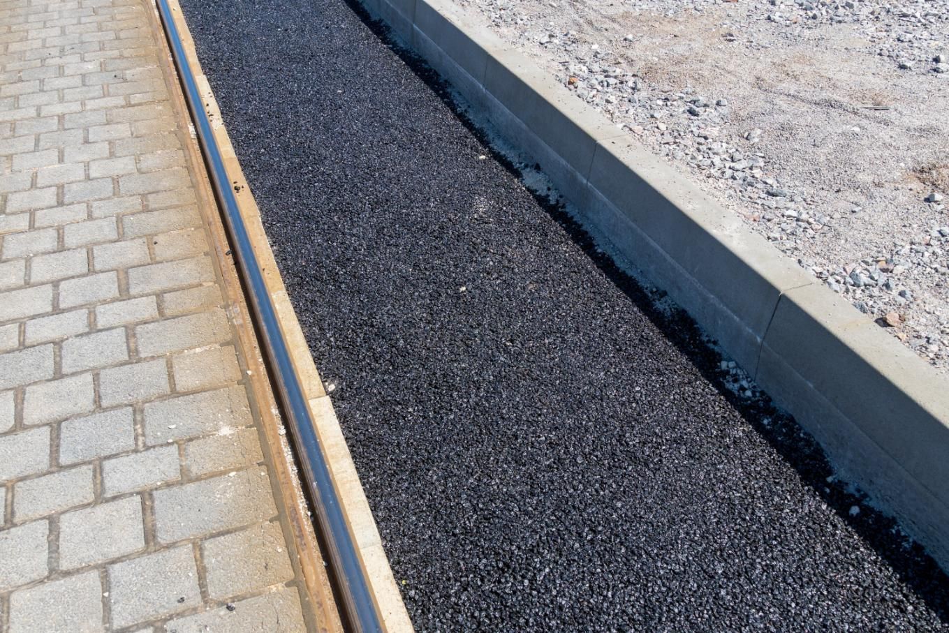 A paved path featuring a strip of asphalt bordered by gray stone pavers and a concrete curb on a construction site.