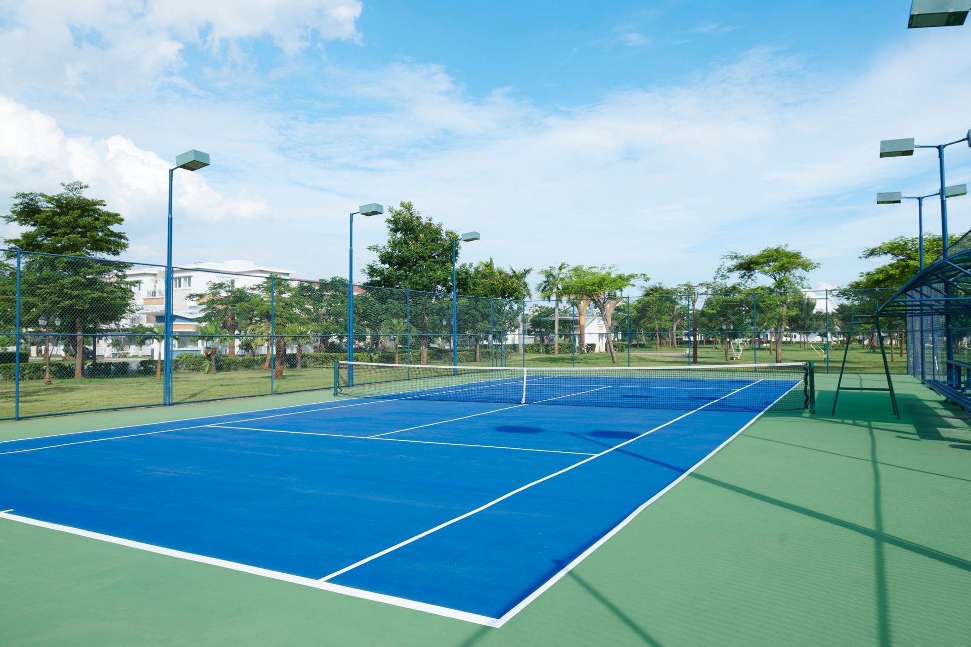A vibrant blue pickleball court with white boundary lines on a green surface, surrounded by light poles and green trees.