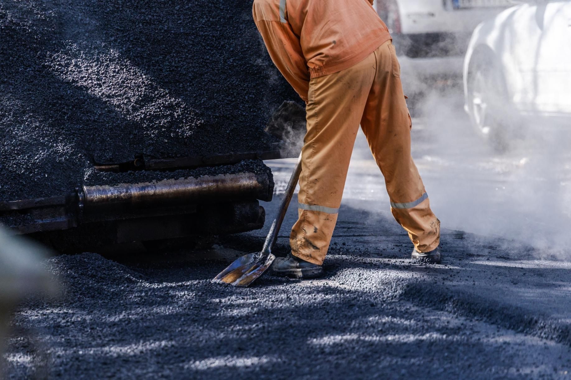 A worker in tan coveralls uses a shovel to spread hot, steaming asphalt on a road.