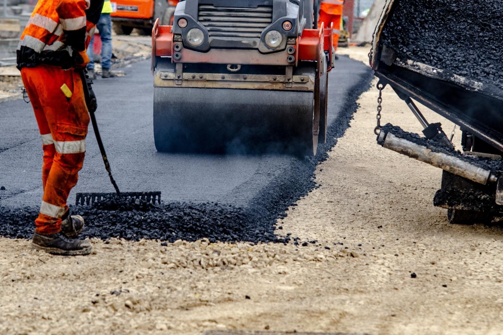 A construction worker in high-visibility gear spreads asphalt while a road roller flattens the surface at a job site.