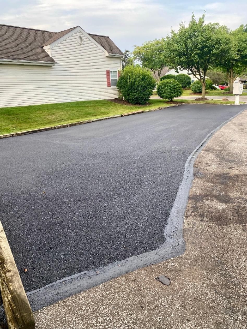 A newly paved dark asphalt driveway meets an older, textured concrete surface in front of a white house with a green lawn.