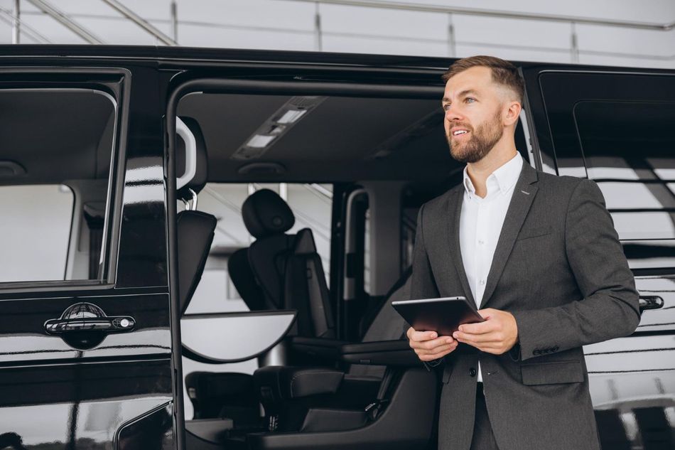 Man in suit holding a tablet, standing by the open door of a black van.