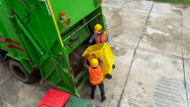 Garbage truck with two workers in safety vests emptying a yellow bin. Concrete, green truck, orange vests.