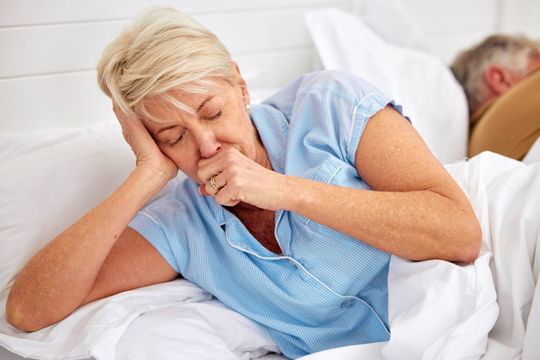 A woman is coughing while laying in bed next to a man who is sleeping.