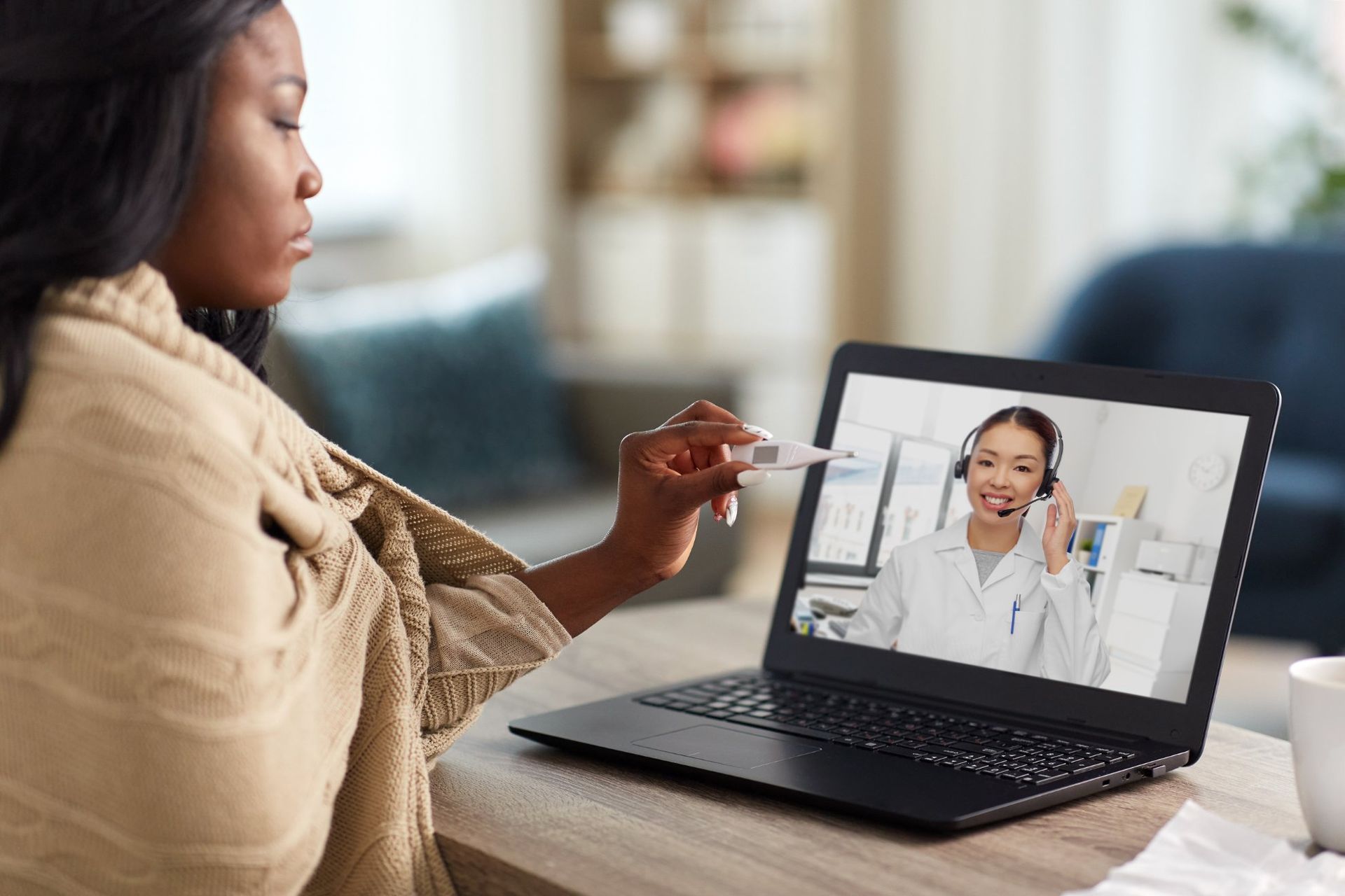 A woman is sitting at a table holding a thermometer in front of a laptop computer.