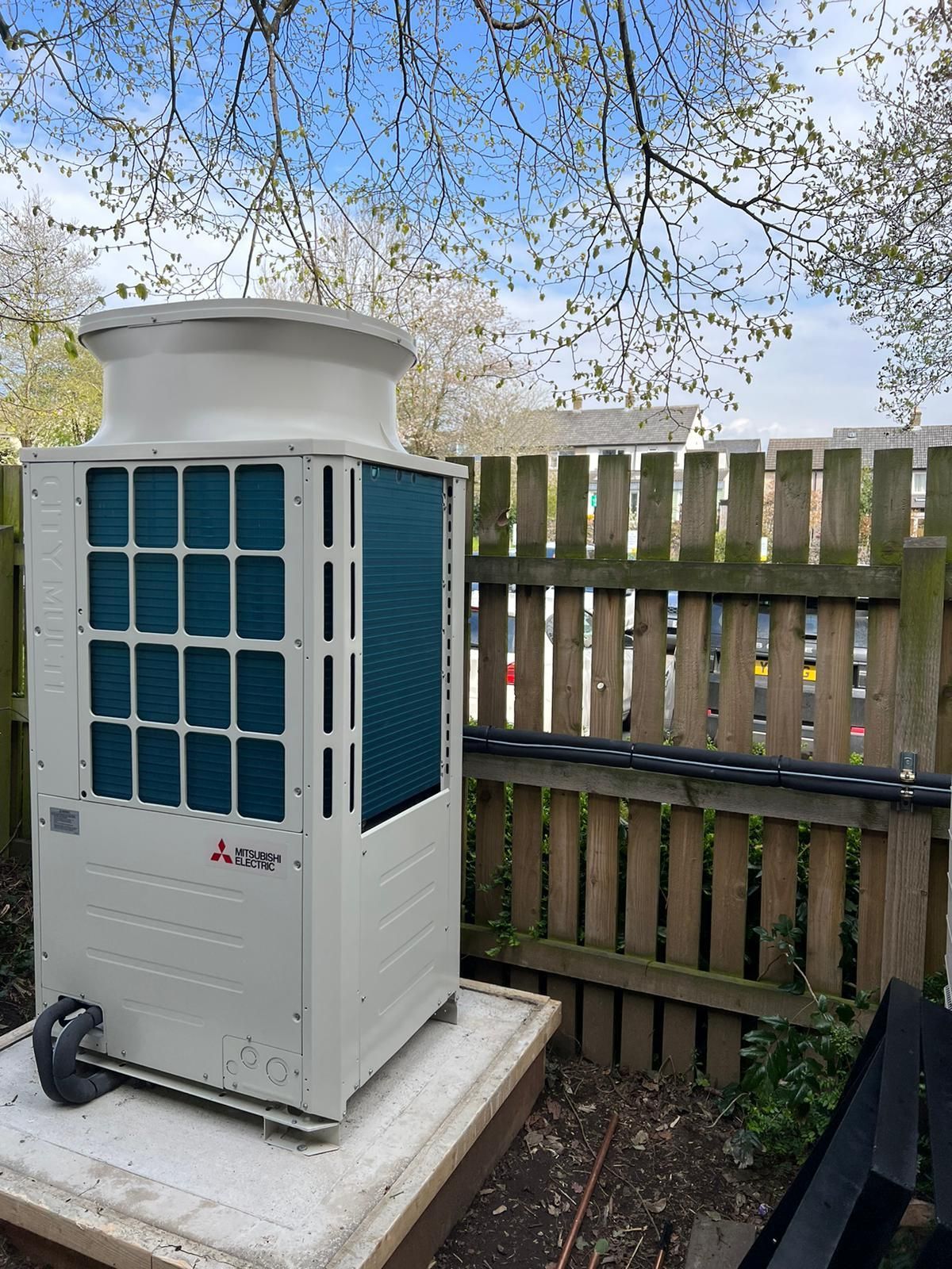 Outdoor HVAC unit on a concrete pad beside a wooden fence, with trees overhead.