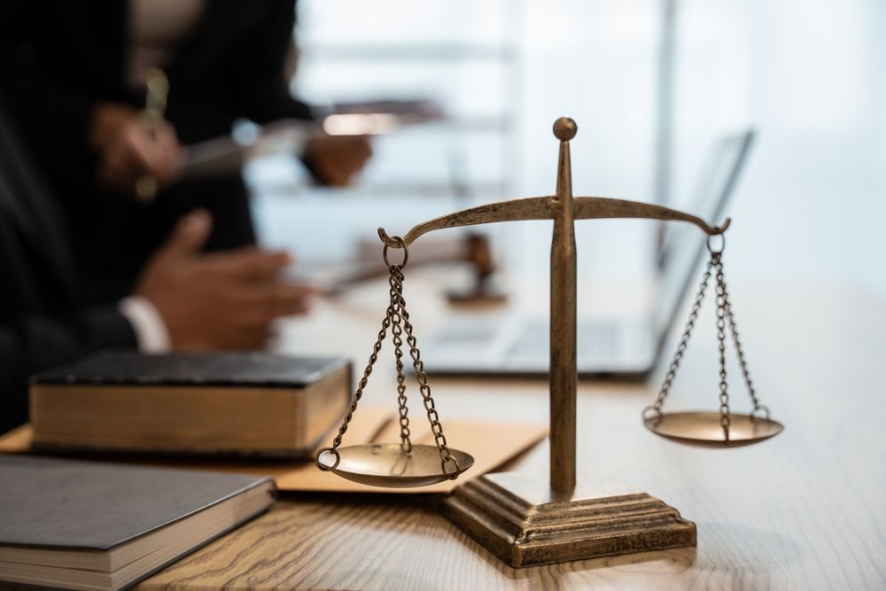 Bronze scales of justice on a desk with law books, a laptop, and people in the blurred background.