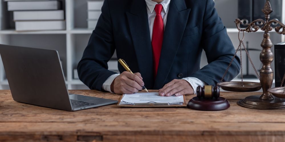A person in a suit signing documents at a desk, with a laptop, gavel, and scales of justice.