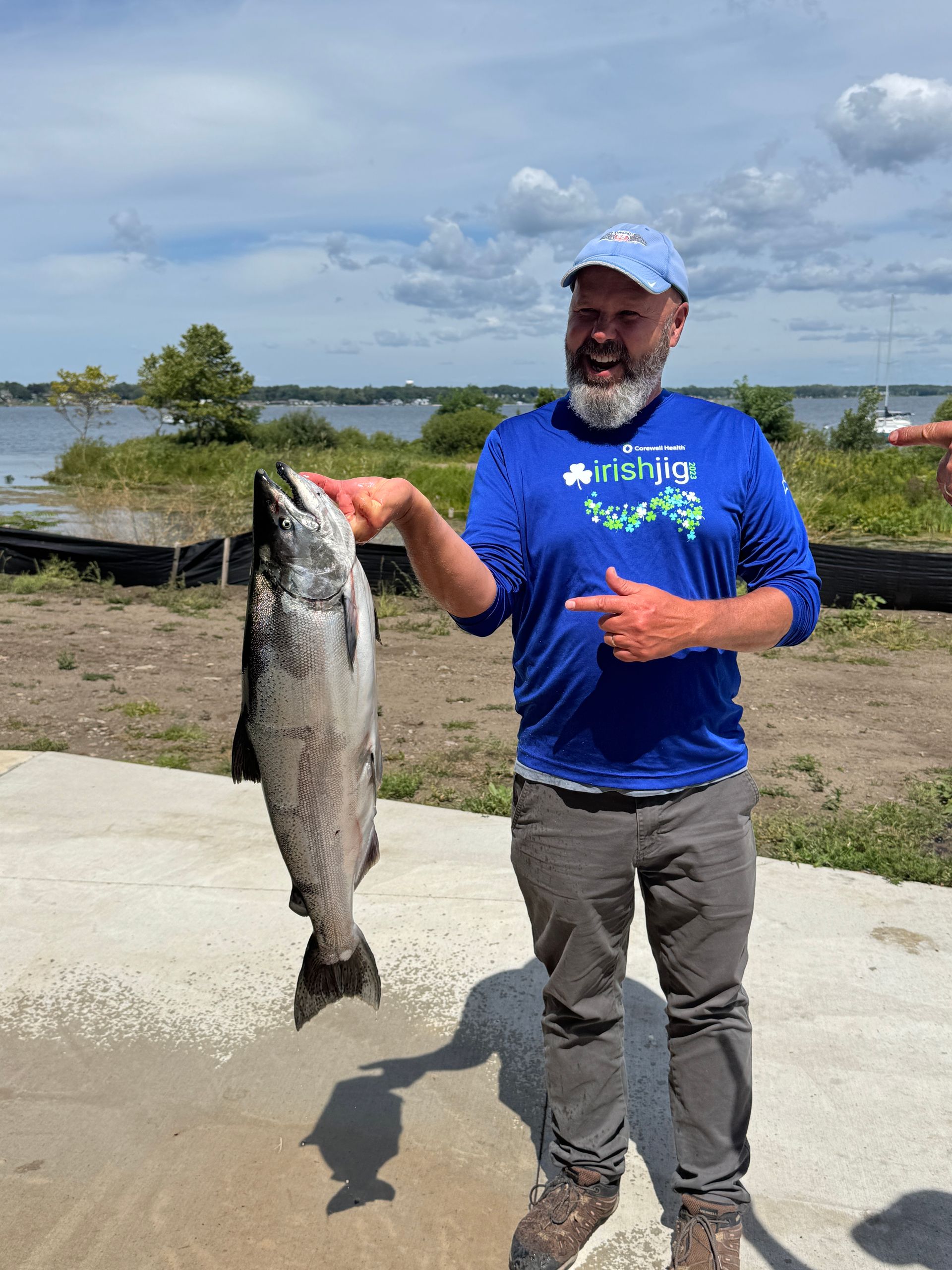 Man in blue points at a large salmon he is holding; sunny outdoor setting with water and sky.