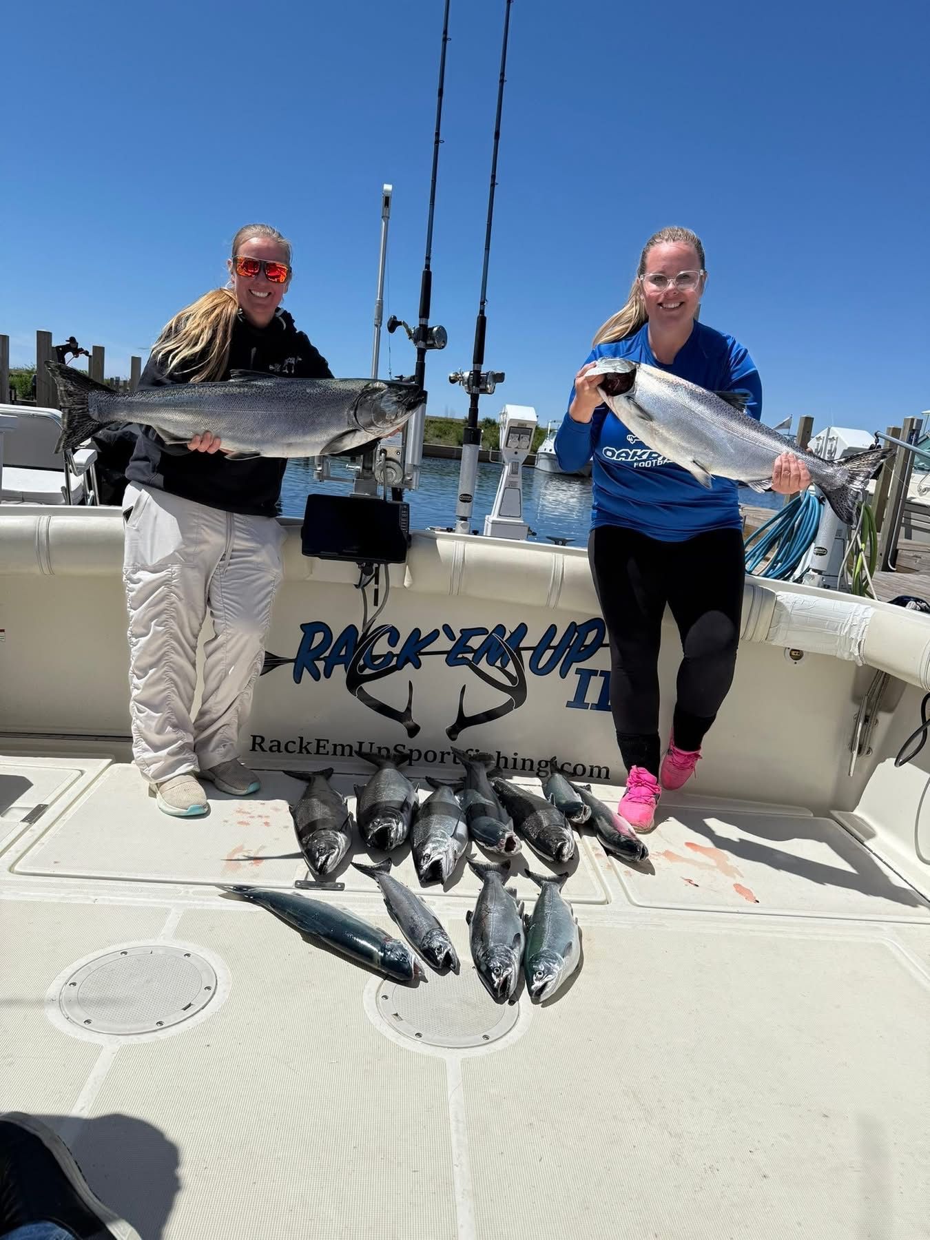 Two people holding fish on a boat; various fish are on the deck. Blue sky, sunny day.