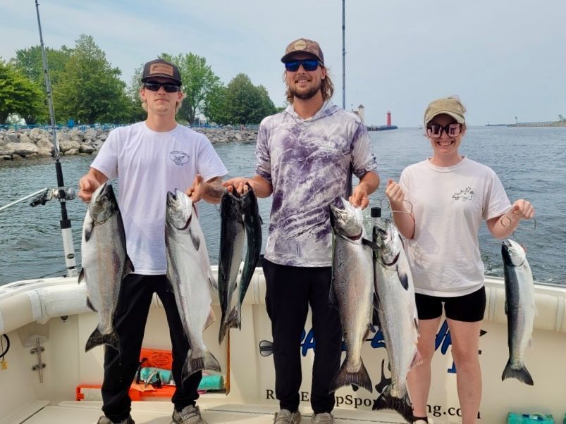Three people on a boat display several large fish they caught; lake in background.