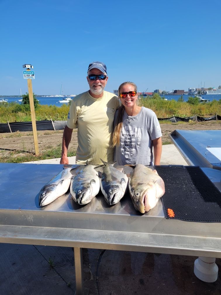 Man and woman standing behind fish on a metal table, with boats and blue sky in the background.