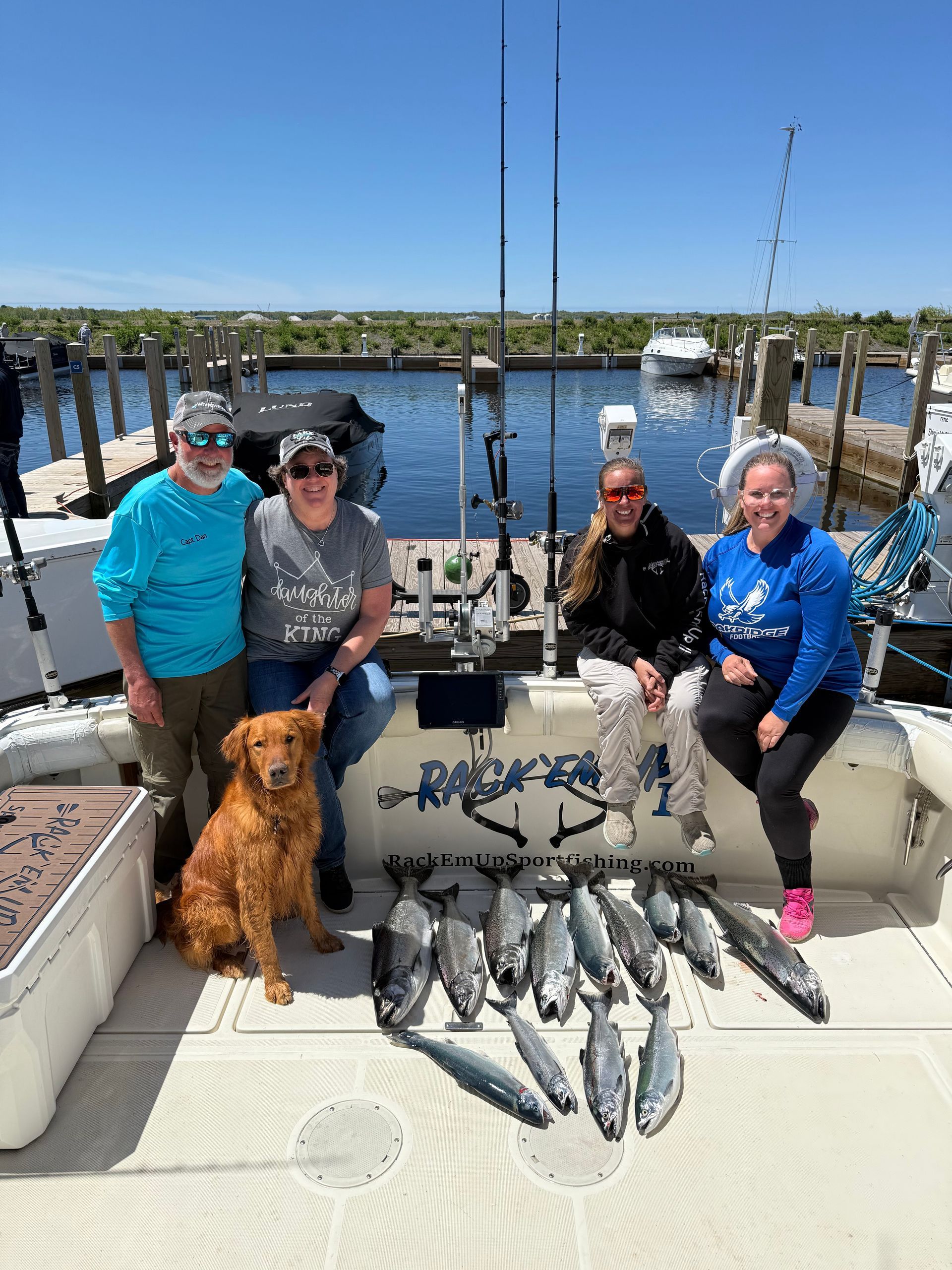 Group and dog pose with caught fish on a boat; sunny day, harbor in background.