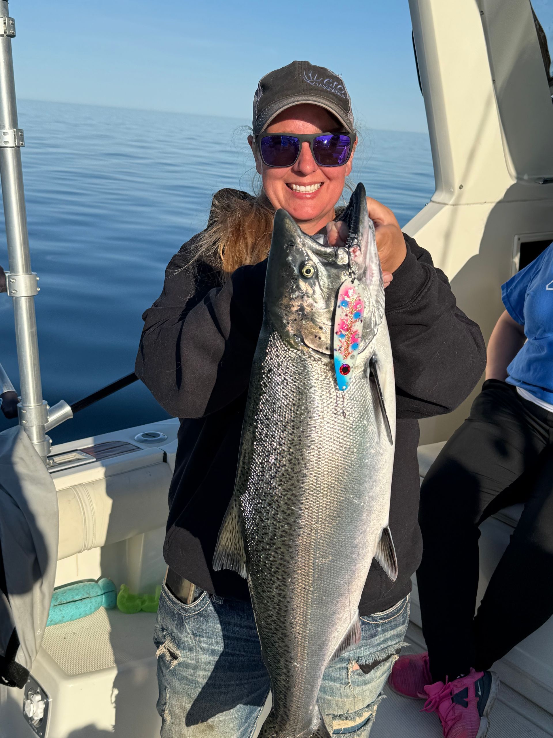 Woman on a boat holding a large, silver fish she caught; smiling. Blue water and sky in the background.