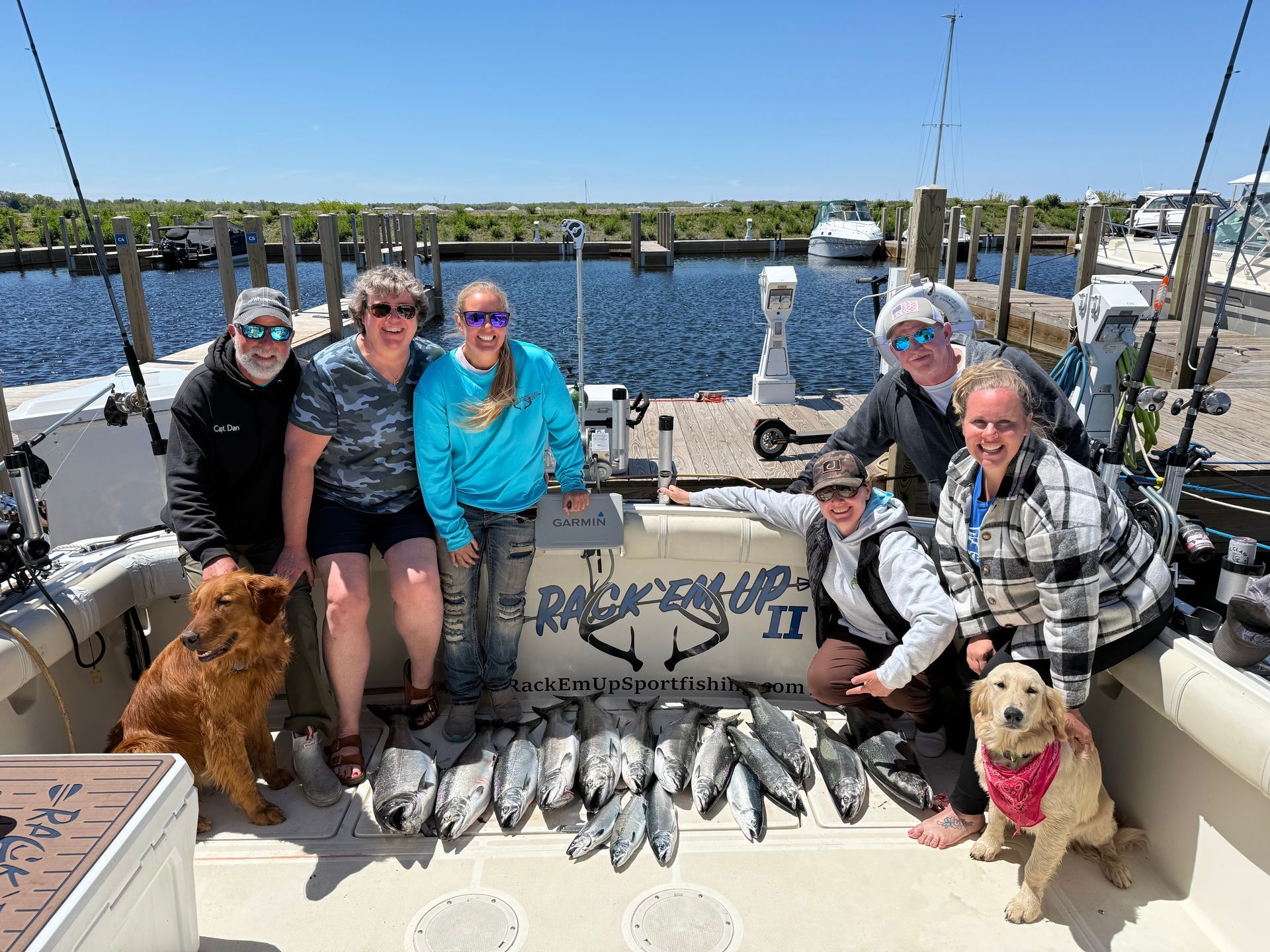 Group of people on a boat with caught fish and two dogs, posing at a dock on a sunny day.