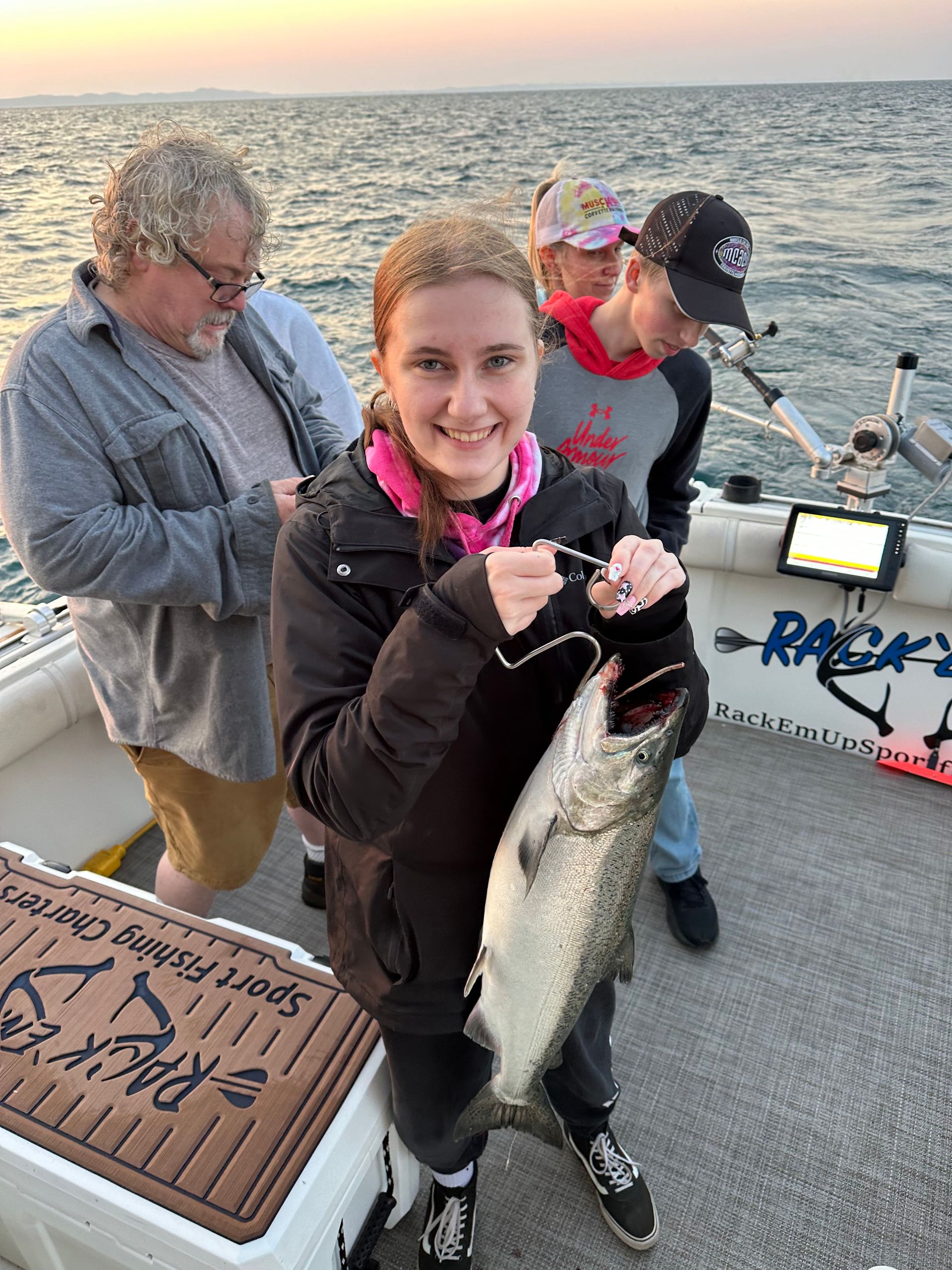 Girl smiles holding a fish on a boat, two people watch. Setting is a body of water with a sunset.