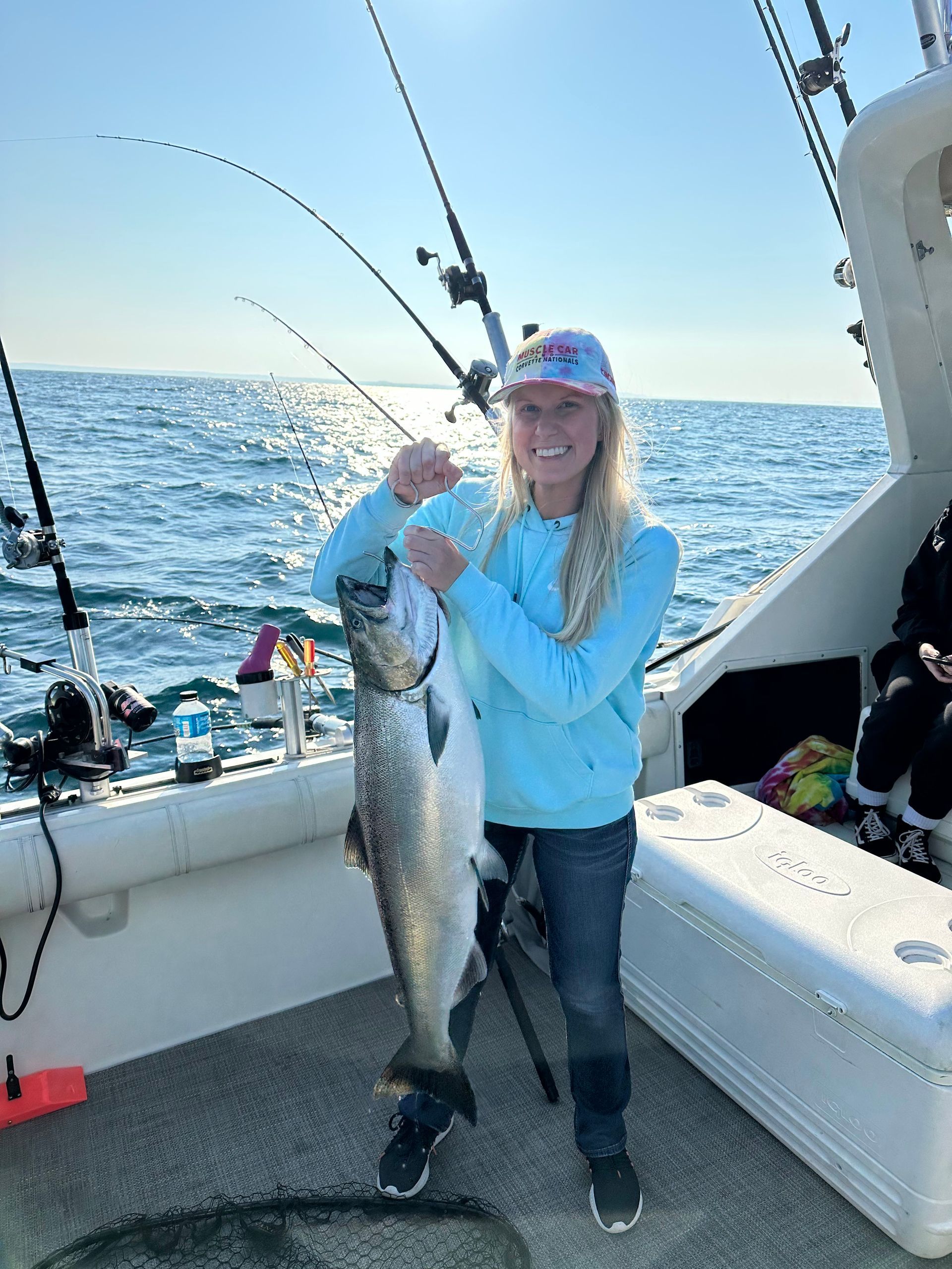 Woman on a boat smiles while holding a large fish; blue water and rods in the background.