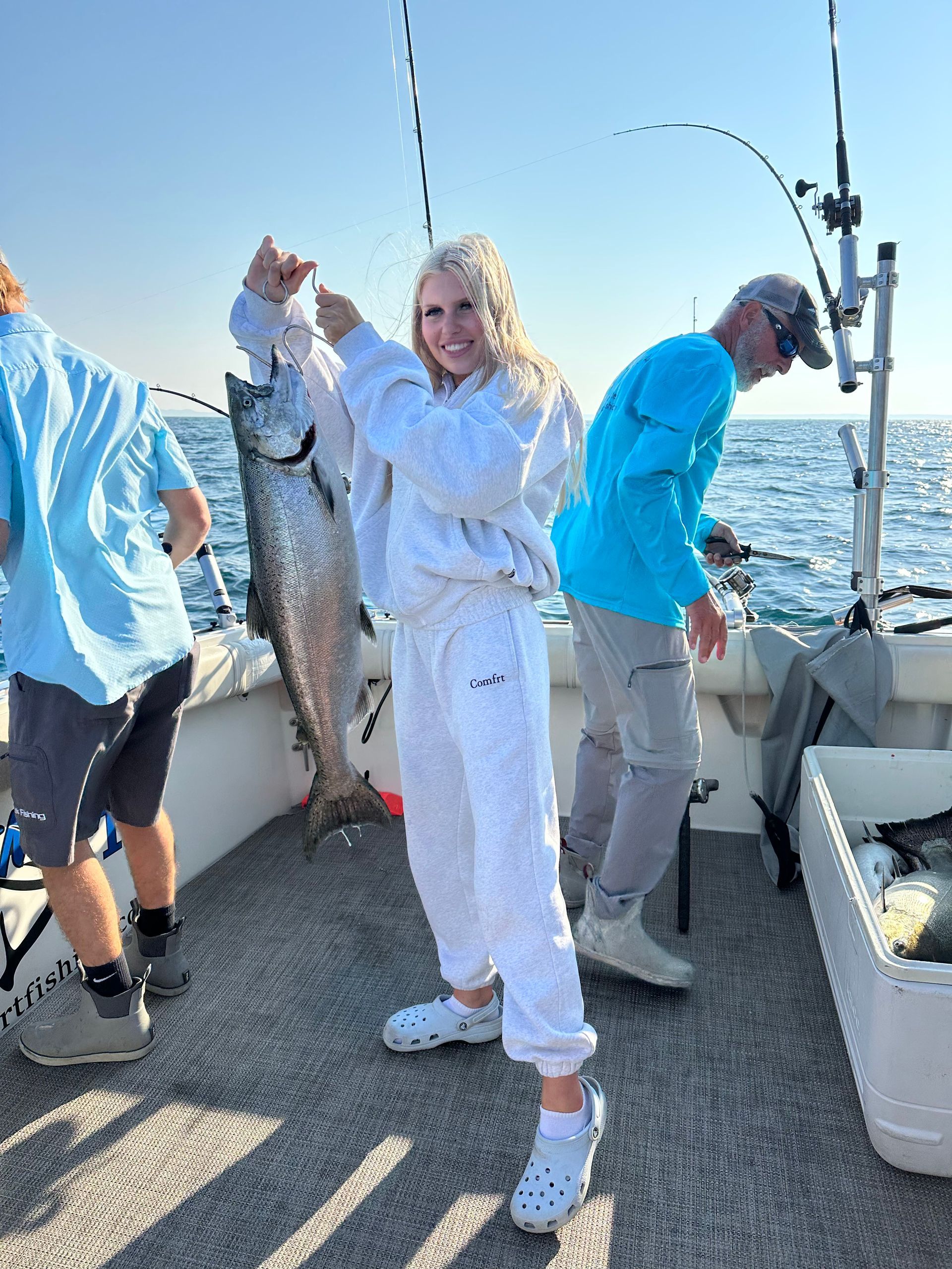 Woman holding up a large fish on a boat; two other people fishing. Blue water and sky.
