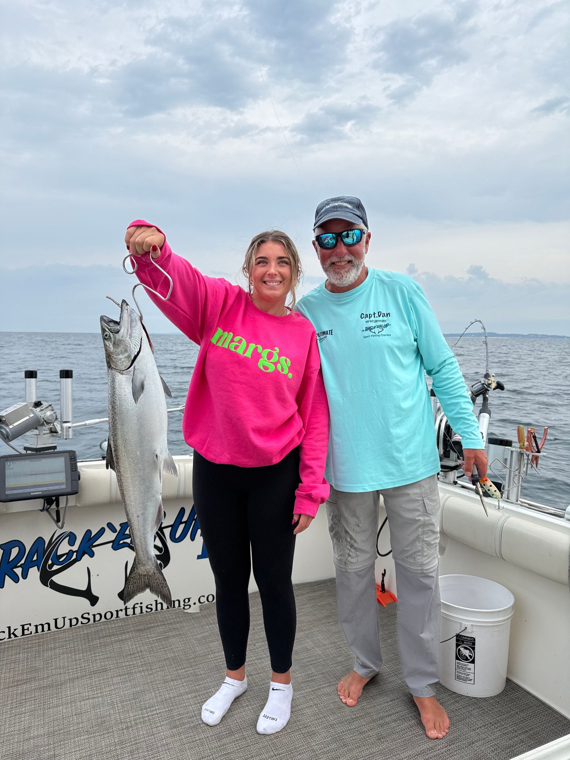 Woman in pink sweatshirt and man holding a fish on a boat. Cloudy sky, fishing gear visible.