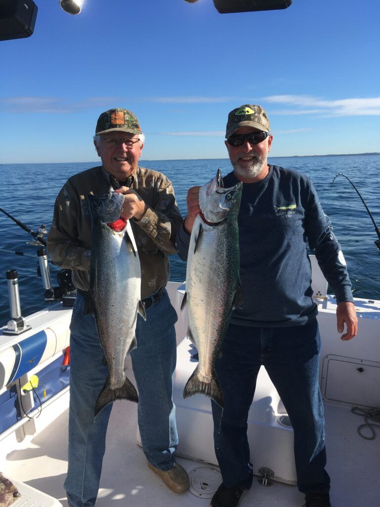 Two men on a boat holding large silver fish. Blue sky, water visible.
