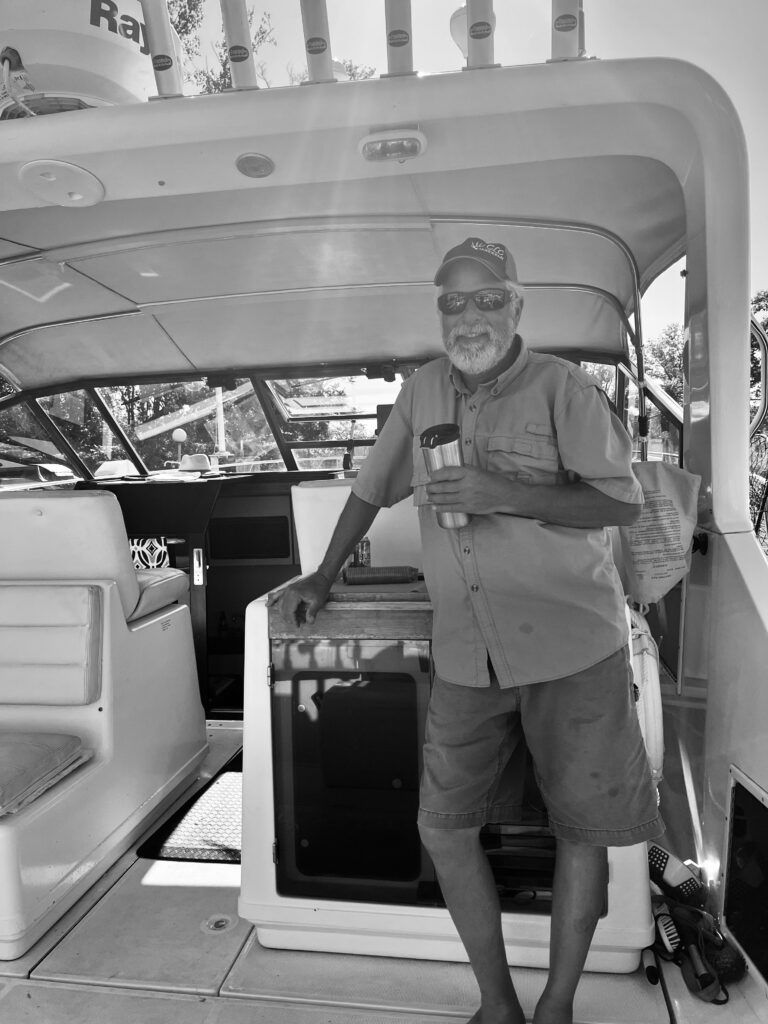 Man standing on a boat, wearing a hat, sunglasses, and shorts, holding a cup.