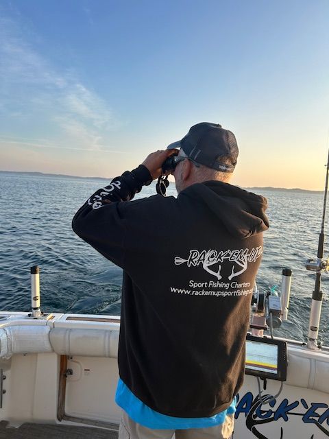 Man on a boat looking through binoculars at the ocean. Wearing black hoodie, blue shirt, cap.