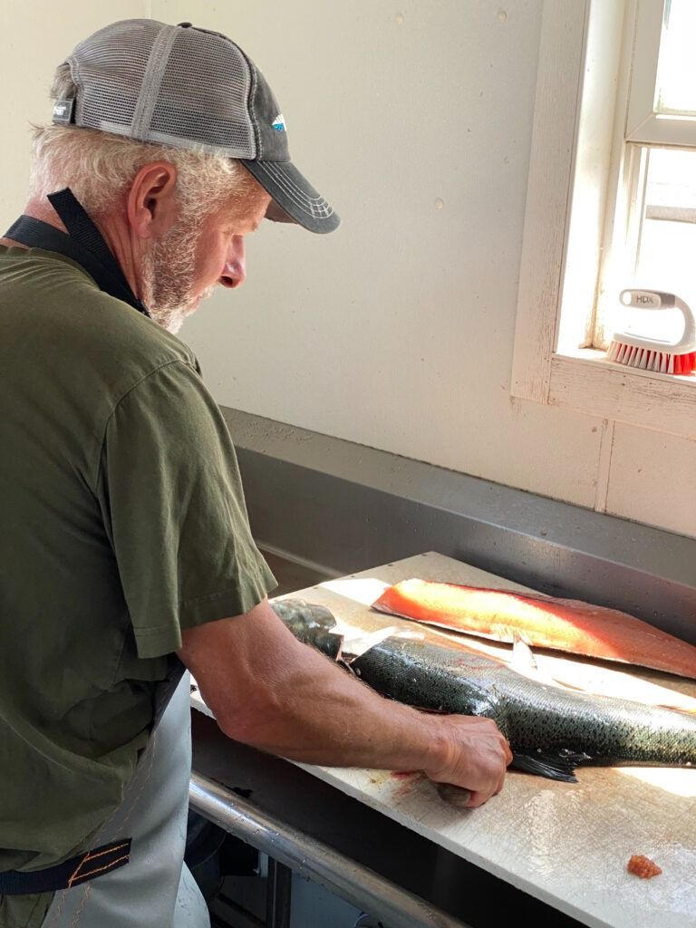 Man in hat fillets a fish at a workstation, salmon fillet on the counter.