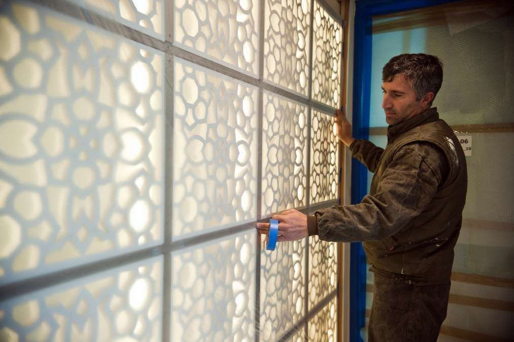 Man installing an ornamental panel with a floral design. He's wearing a jacket, in a workshop setting.