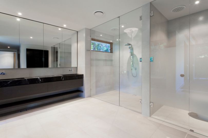 Modern bathroom with large glass shower, white floors, dark vanity, and mirrored walls.