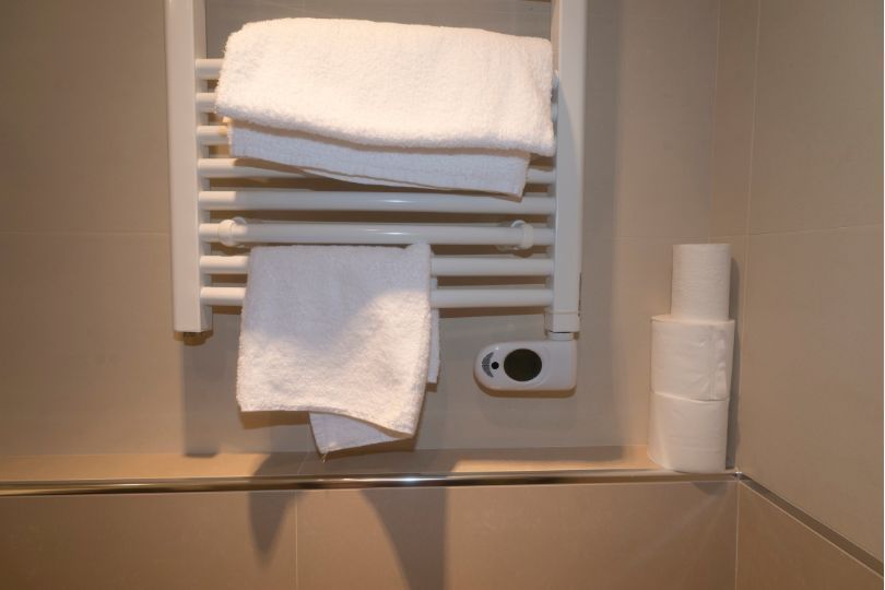 White towels on a heated rack in a bathroom next to toilet paper rolls.