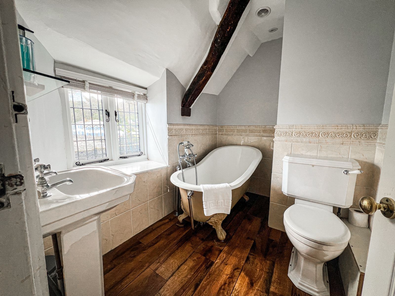 Bathroom with a claw-foot tub, toilet, and sink, under a slanted ceiling. Wooden floor and tan tile walls.
