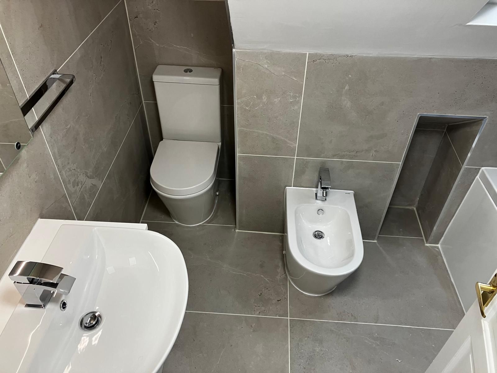 Small gray-tiled bathroom with toilet, sink, bidet, and shower area.