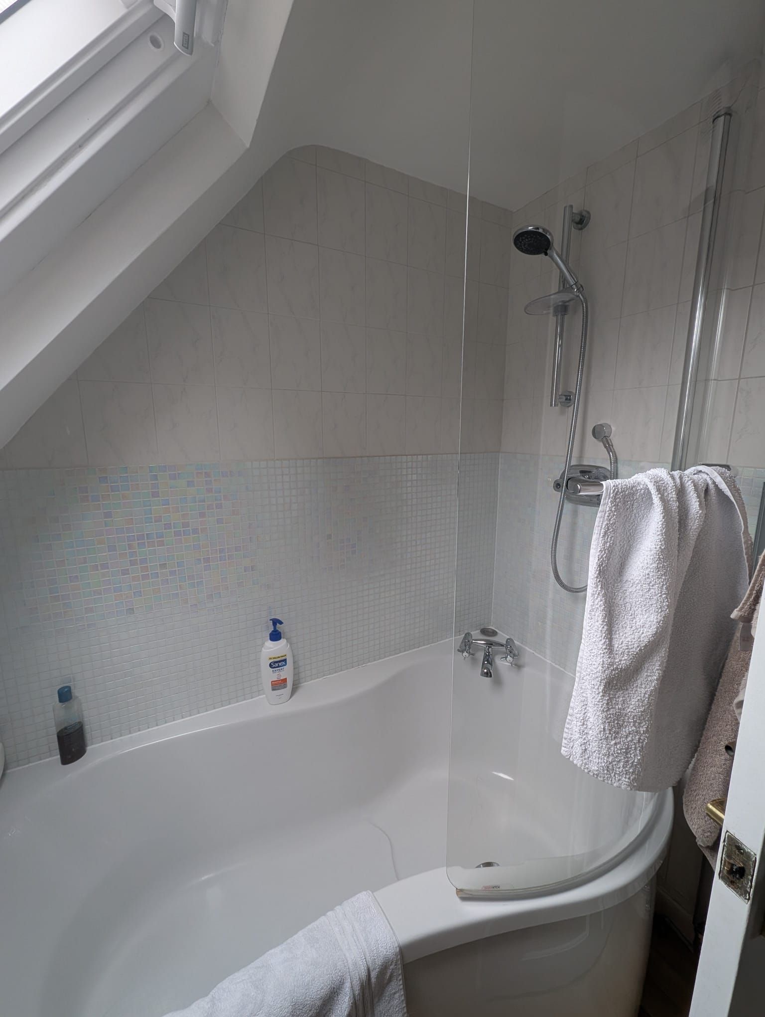 Bathroom with a white bathtub, shower, and angled ceiling; white and gray tiles; white towels.