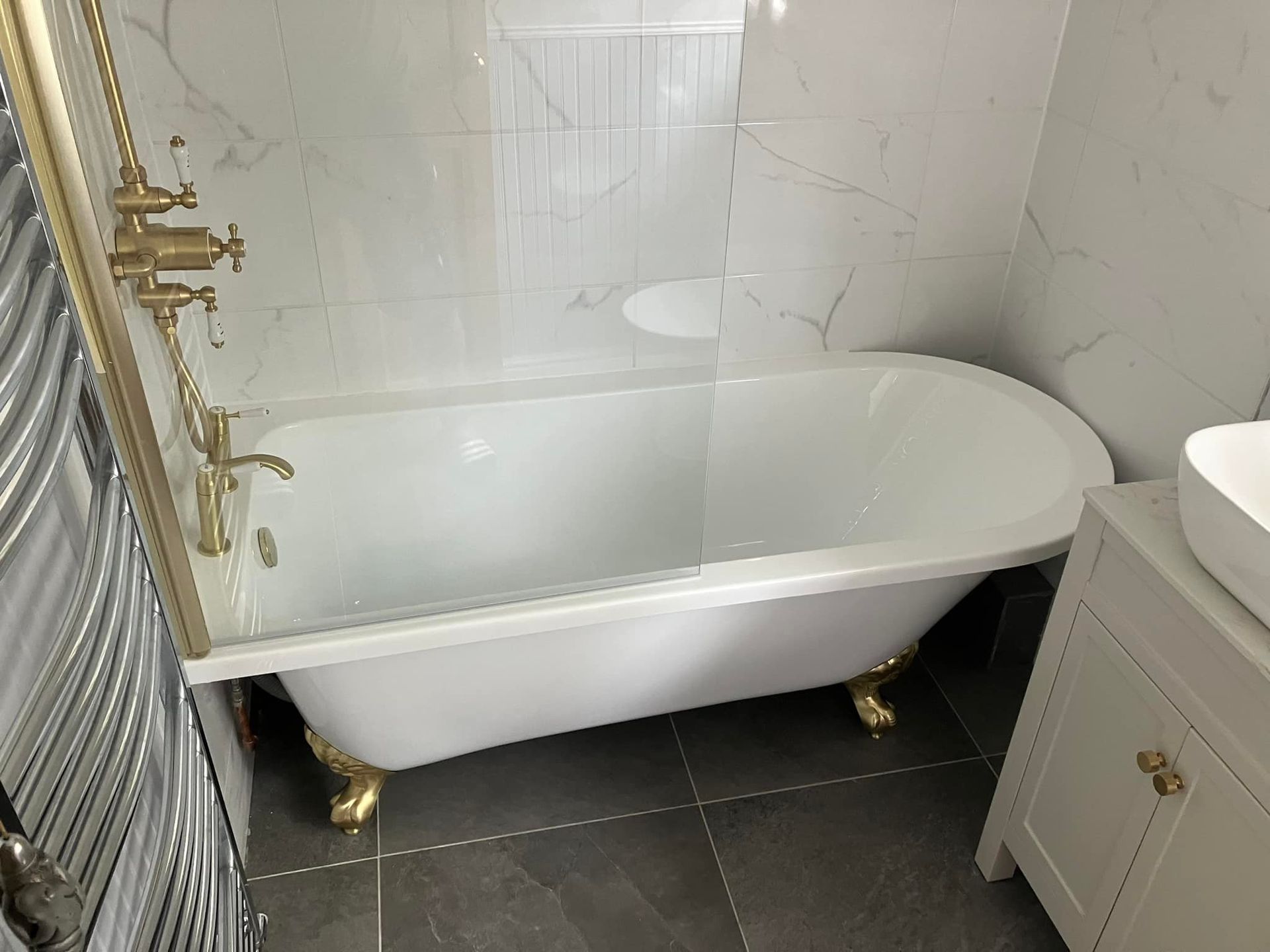 Claw-foot bathtub with gold fixtures in a modern bathroom with marble-look tiles and a white vanity.