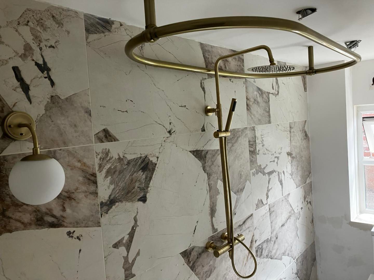 Bathroom with marble-patterned tiles, brass shower fixtures, and a round light fixture.