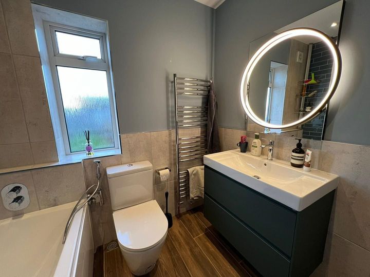 Bathroom with toilet, sink, and towel warmer; blue-green vanity, round illuminated mirror, neutral walls.