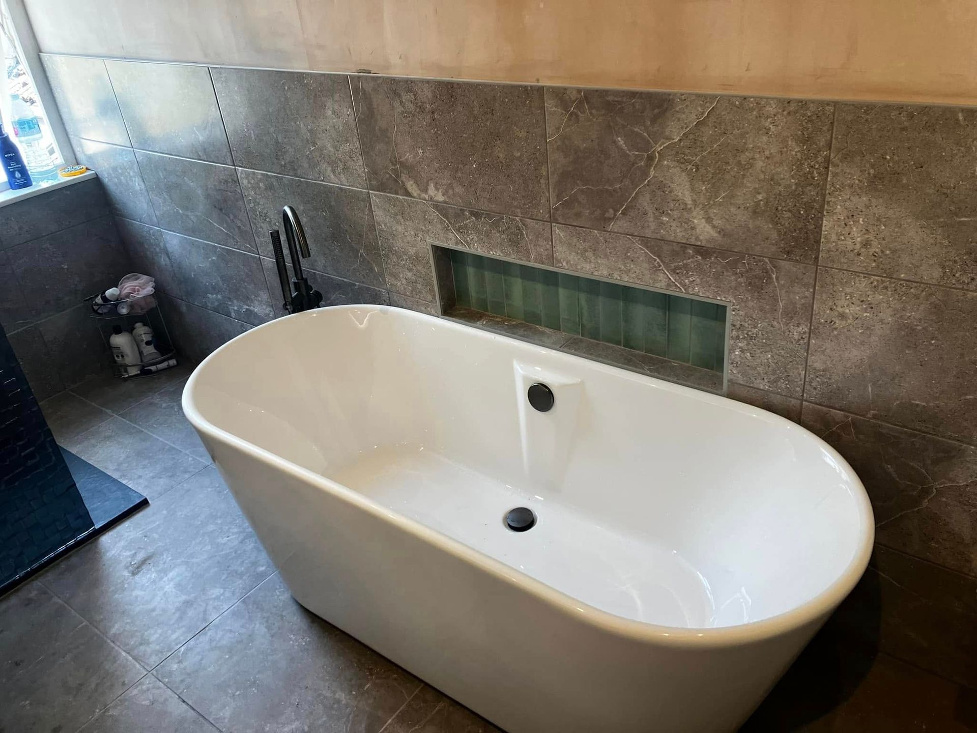 White oval bathtub against a grey tiled wall with a recessed shelf.