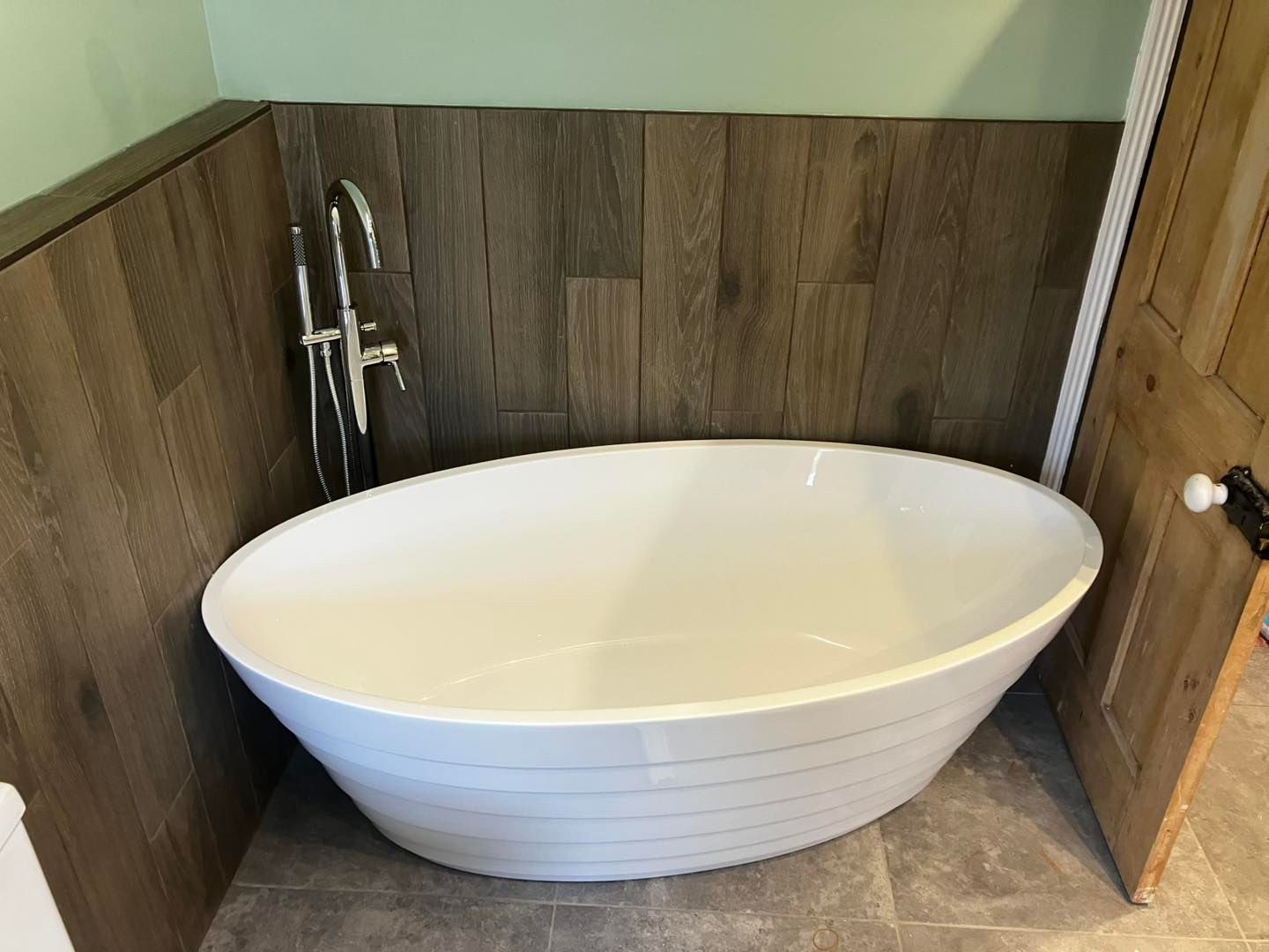 White oval bathtub against wood-paneled wall with a faucet and partially open wooden door.