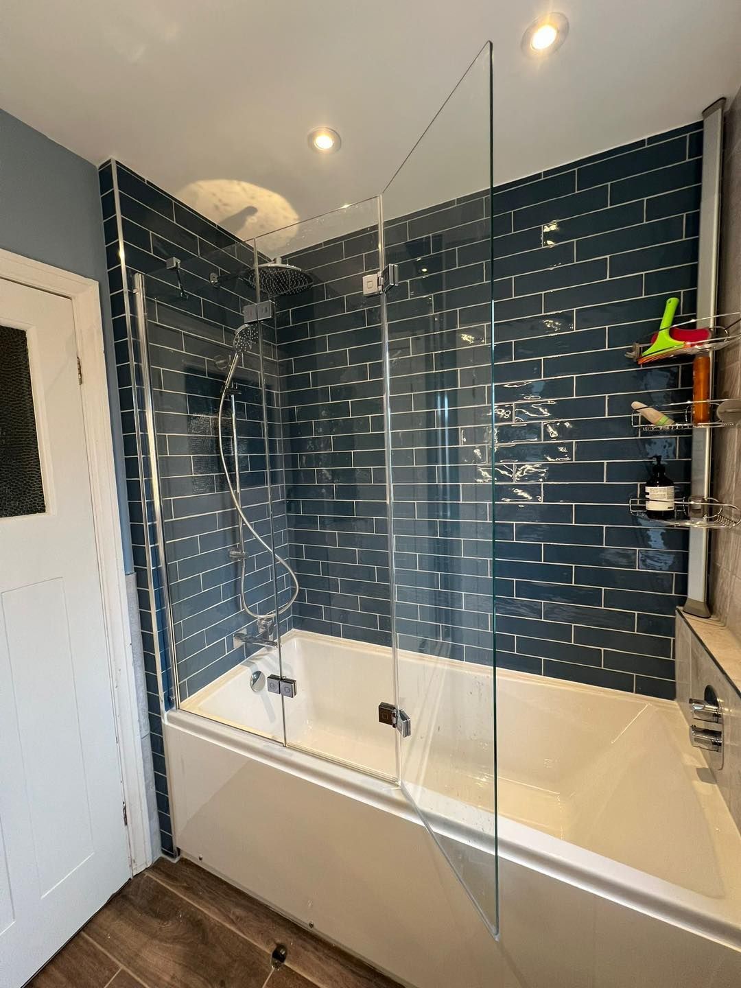 Blue-tiled shower enclosure with glass screen and chrome fixtures. White tub against a blue wall.